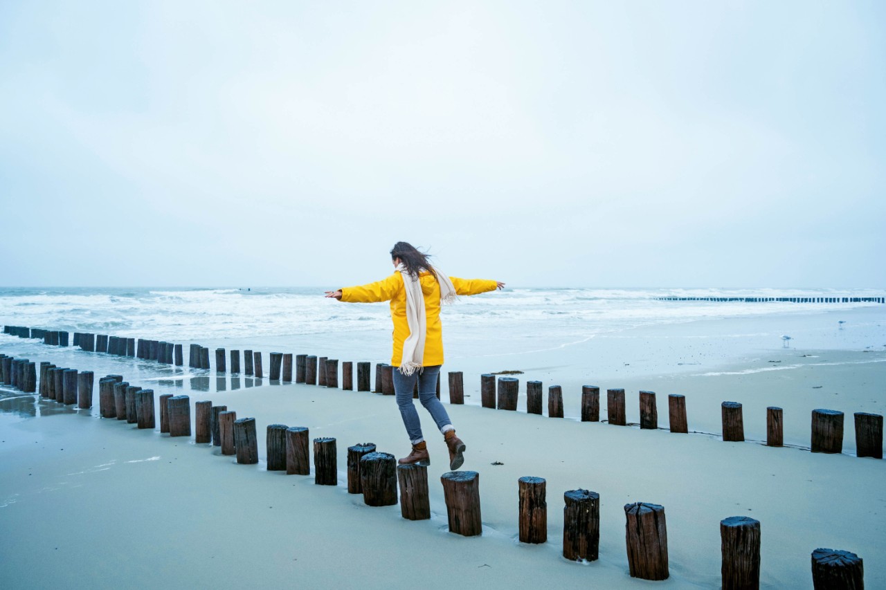 Vrouw loopt over paaltjes op strand in gele jas