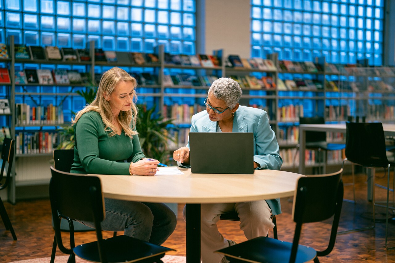 Twee vrouwen zitten aan tafel in bibliotheek met laptop