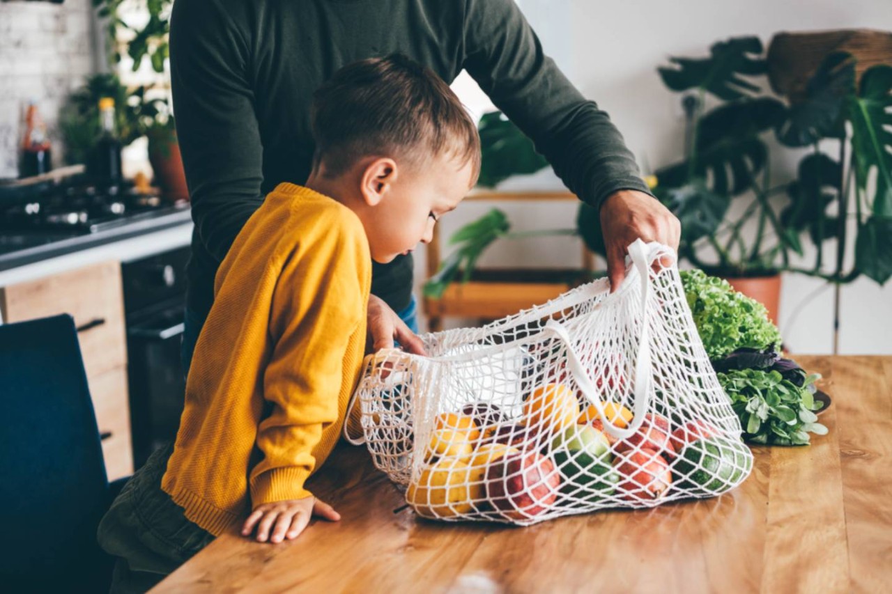 Jongetje met gele trui kijkt in een boodschappennet met fruit op tafel
