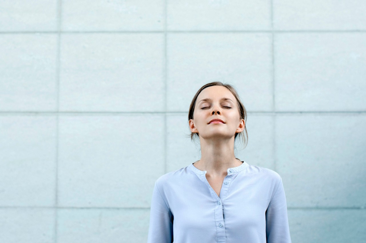 Relaxed pretty young woman standing at wall outdoors. Female student or business woman relaxing with her eyes closed. Serenity concept. Front closeup view.