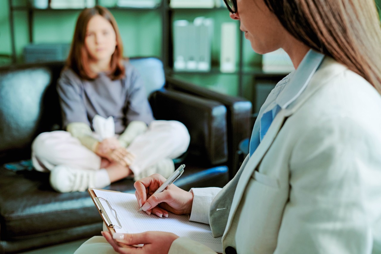 Therapist wearing suit taking notes while counseling young woman sitting on couch in office providing professional support and advice for client's mental health