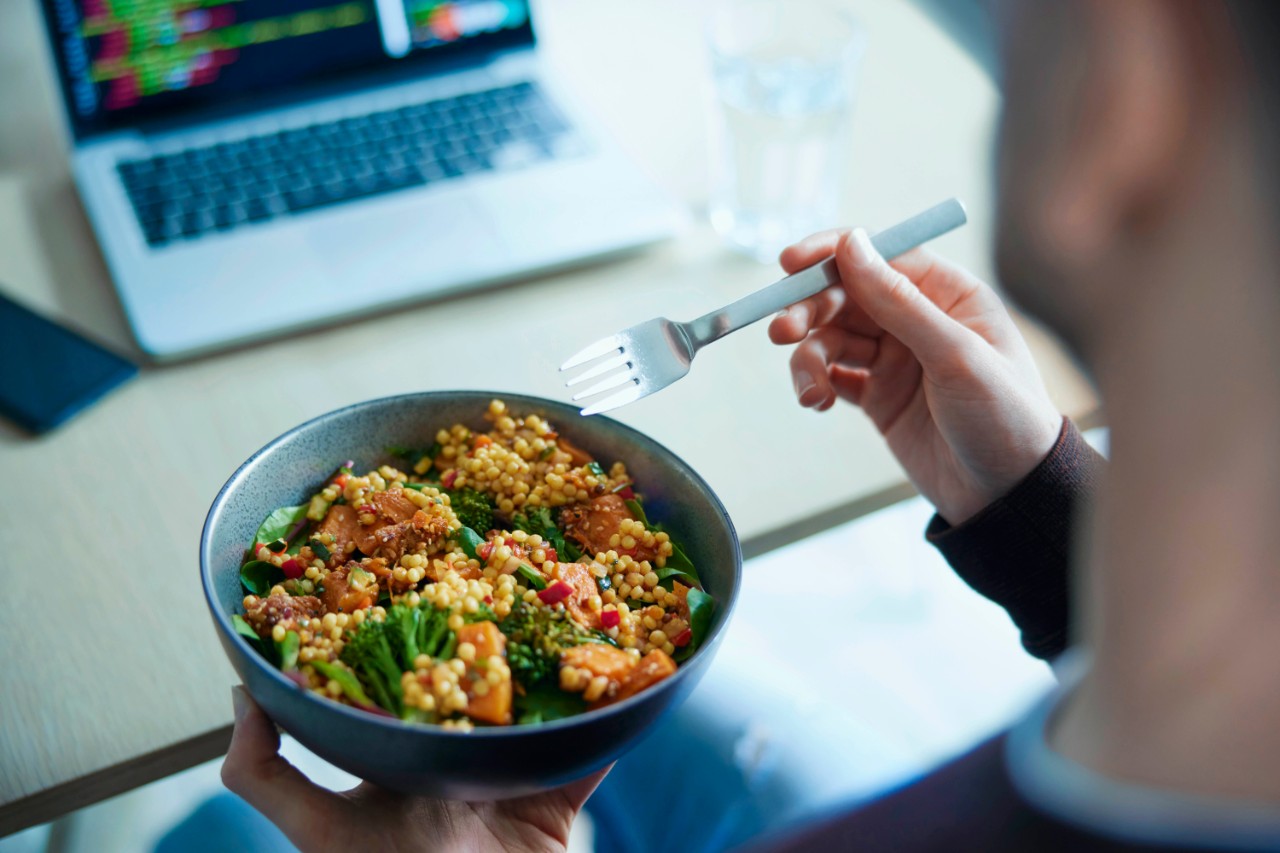 Close Of Man Eating Healthy Vegan Lunch At Office Deak Whilst Using Laptop