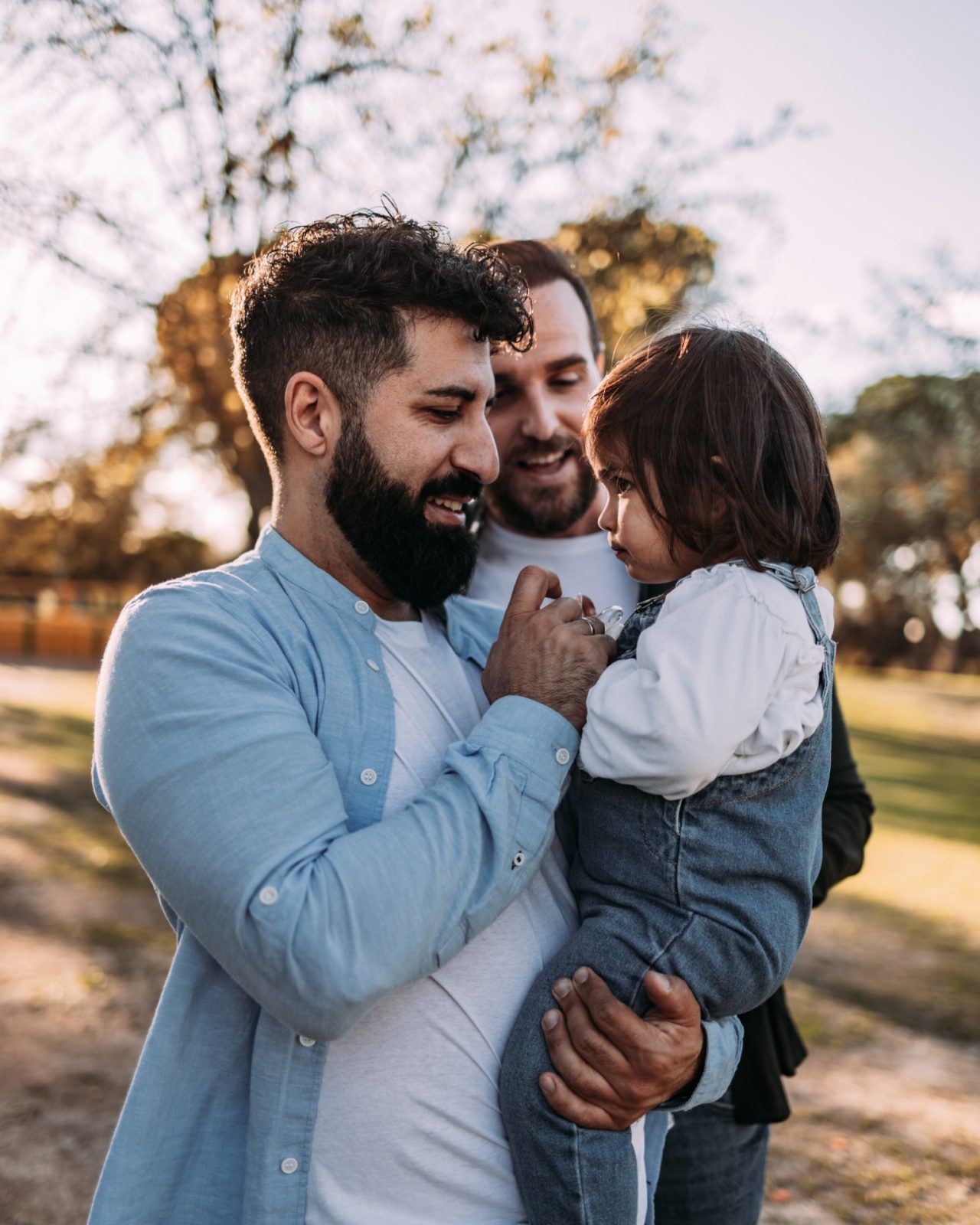 Happy male gay couple with their daughter in their arms. Her father gives her the pacifier.