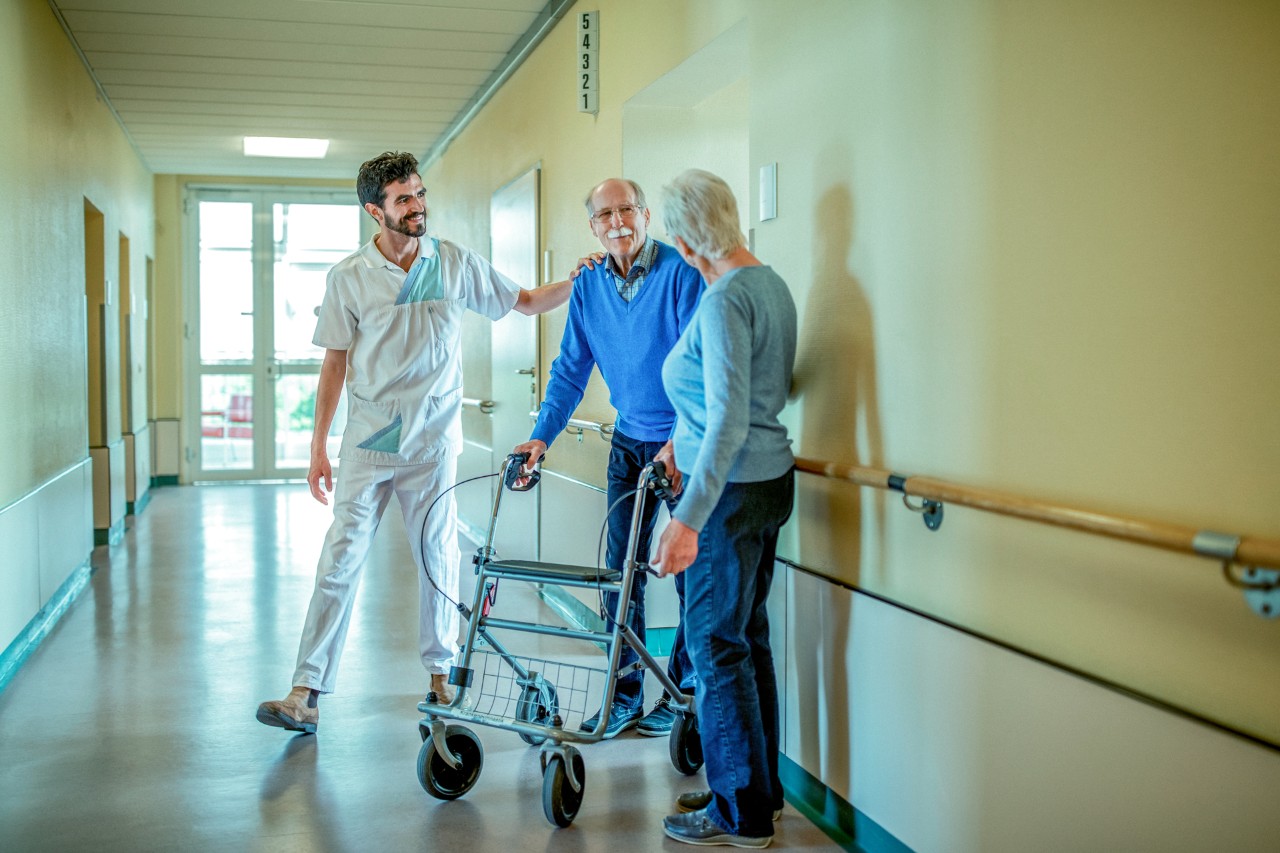 Ouder echtpaar waarvan man met rollator samen met verpleegkundige op gang.